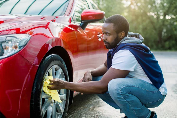 Car washing on open air. Young hipster African bearded man cleaning a  wheel, car rims of modern luxury red car with yellow microfiber cloth, at  outdoor car wash self service - Stock